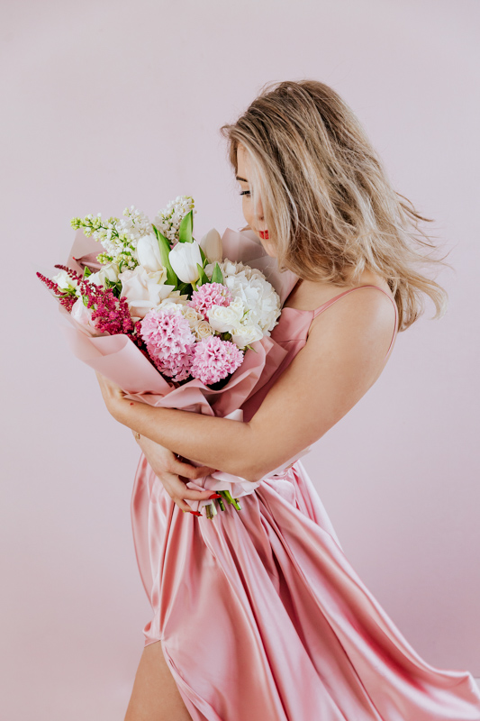 Femeie tanara imbracata in rochie roz in peisaj de iarna care sta pe un fotoliu tine in brate un buchet de flori colorate